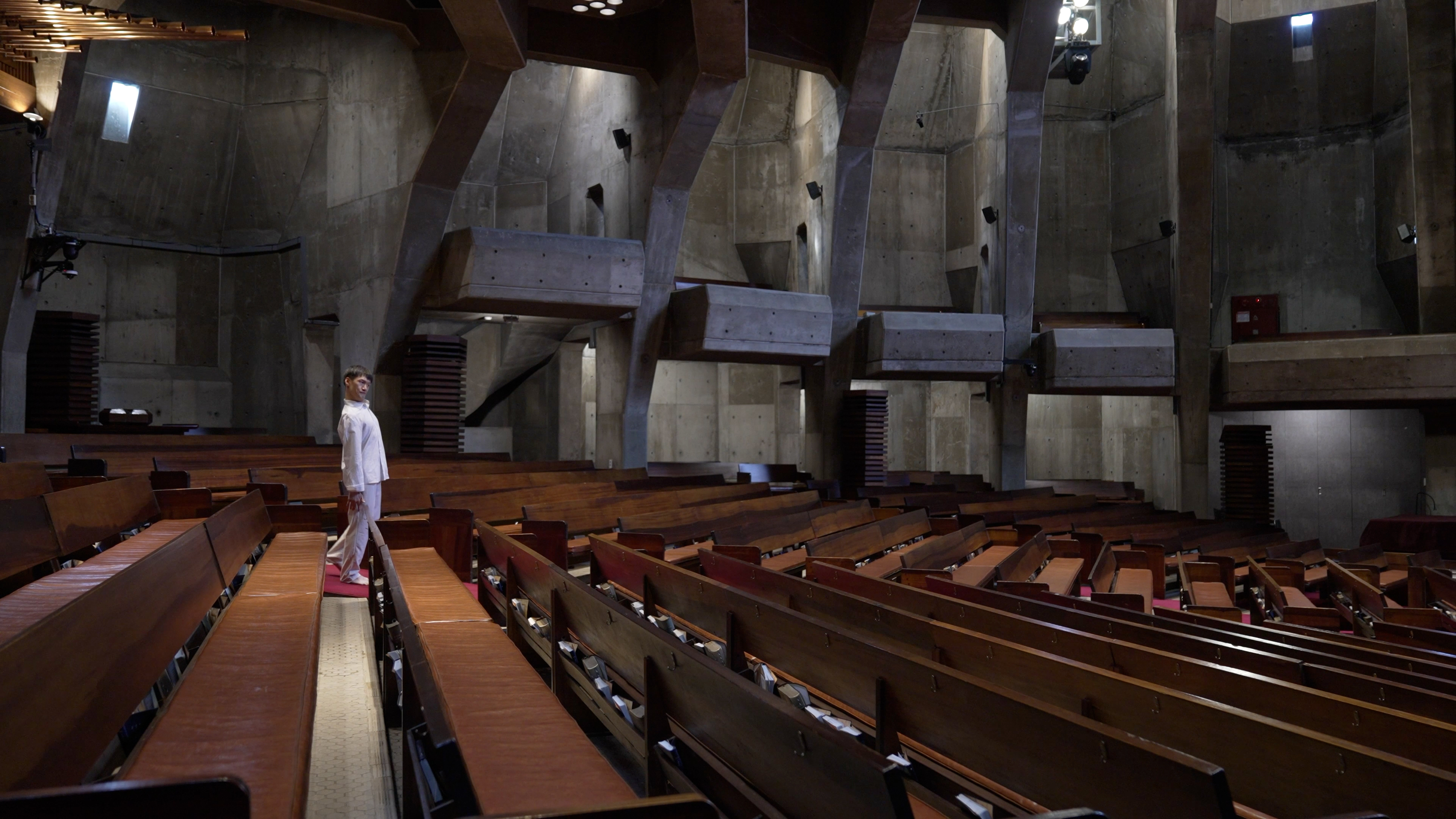 99f9f6d225e55e2c3b859f2a9457eeeb.jpg 문승현 <경계에서> 영상 작품 캡쳐 이미지. 거대한 노출 콘크리트 구조의 경동성당 안에, 갈색 목재 의자들이 줄지어 배열되어 있다. 통로의 끝에는 흰 옷을 입은 문승현 작가가 서 있다.